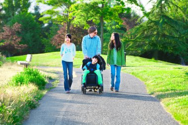 Disabled boy in wheelchair walking with family outdoors on sunny clipart