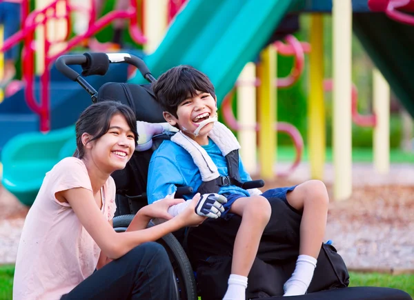 Sister sitting next to disabled brother in wheelchair at playgro — Stock Photo, Image