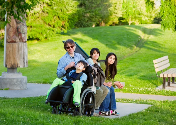 Мальчик-инвалид в инвалидной коляске с семьей на открытом воздухе в солнечный день сидеть — стоковое фото