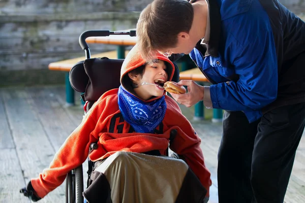 Pai alimentando filho deficiente um hambúrguer em cadeira de rodas. A criança tem Imagens Royalty-Free