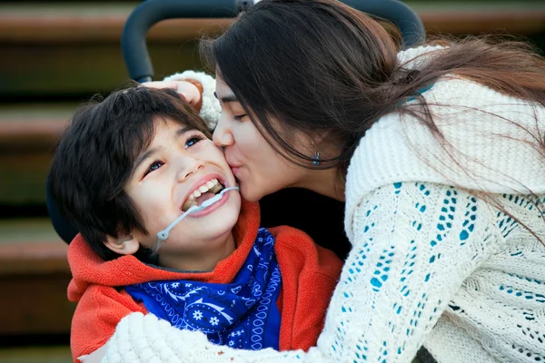 Grande sorella baciare disabili fratellino seduto in sedia a rotelle — Foto Stock