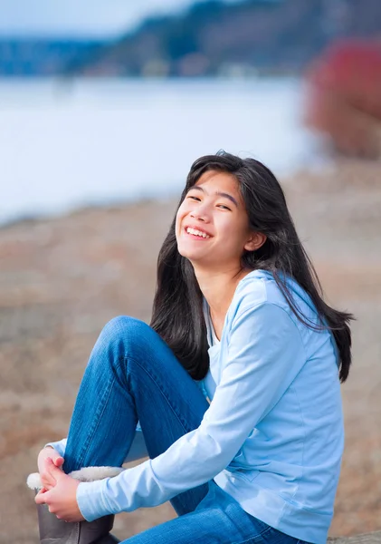 年轻女孩穿着蓝色衬衫和牛仔裤坐沿着岩石湖 — 图库照片
