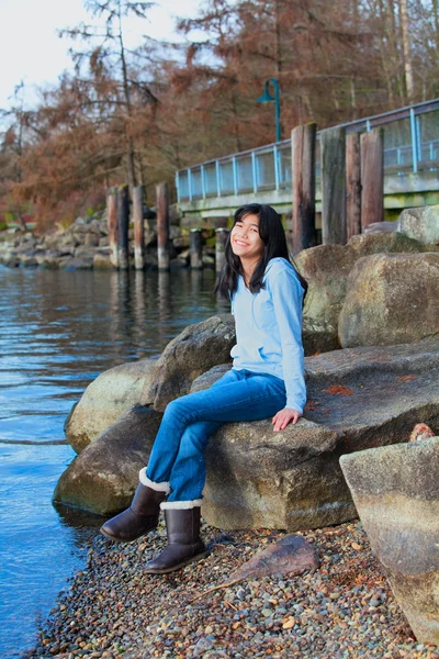 Ung tonåring flicka avkopplande på stora stenblock längs sjöstranden, smil — Stockfoto