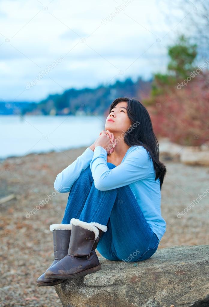 Teen girl sitting on boulder along lake shore praying