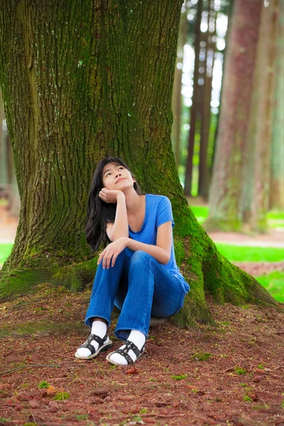 Young teen girl sitting under large pine trees, thinking — Stock fotografie