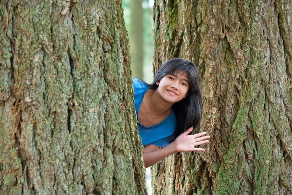 Teen girl peeking through trees, waving and smiling — Stock fotografie