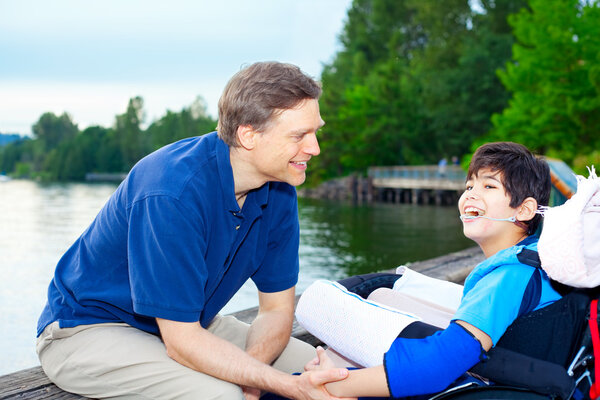 Отец разговаривает с сыном-инвалидом в инвалидной коляске на озере
