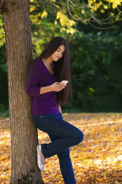 Teen dziewczyna stoi przed jesienne drzewa patrząc na telefon komórkowy — Zdjęcie stockowe