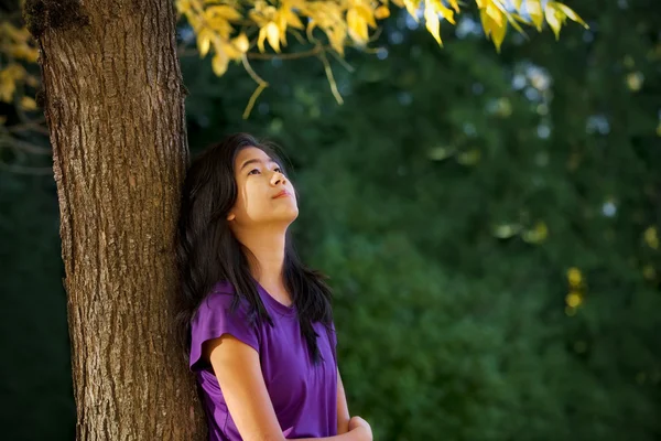Teen girl leaning against tree with autumn leaves looking up — Stock Photo, Image