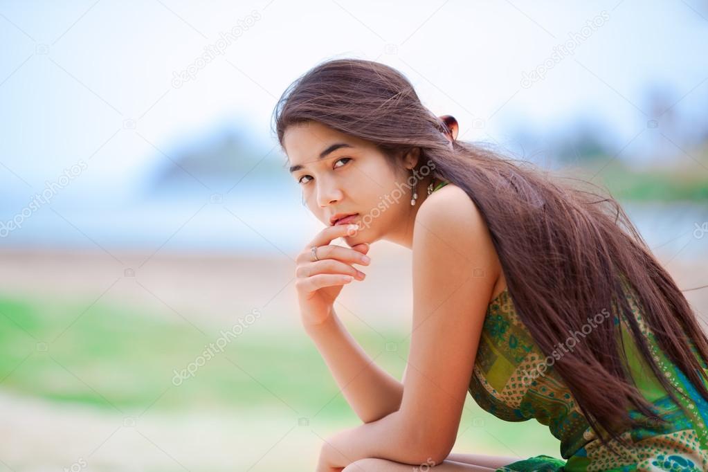 Beautiful biracial teen girl sitting at tropical beach, thinking