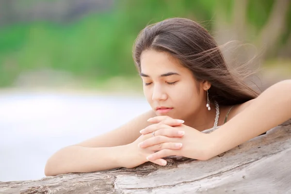 Beautiful teen girl on beach praying by driftwood log — Stock Photo, Image