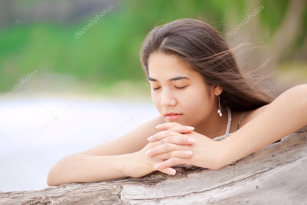 Beautiful teen girl on beach praying by driftwood log 