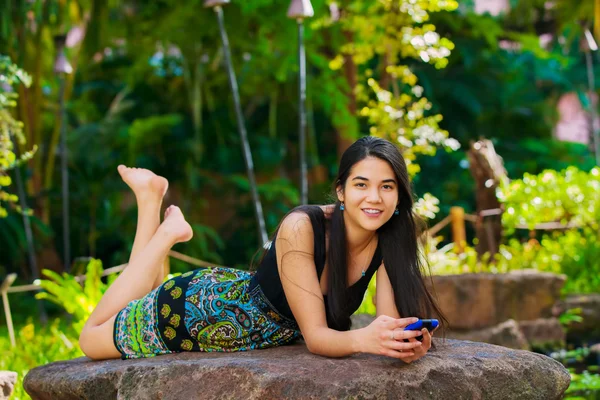 Biracial teen girl lying on rock looking at cellphone outdoors — Stock Photo, Image