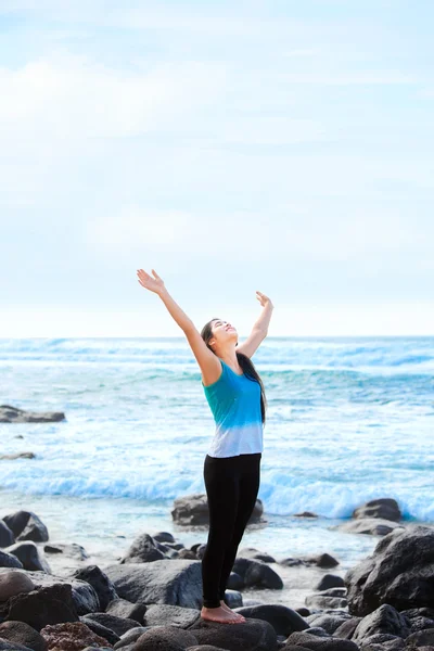 Teen girl standing on rocky beach arms raised, praising God — Stockfoto