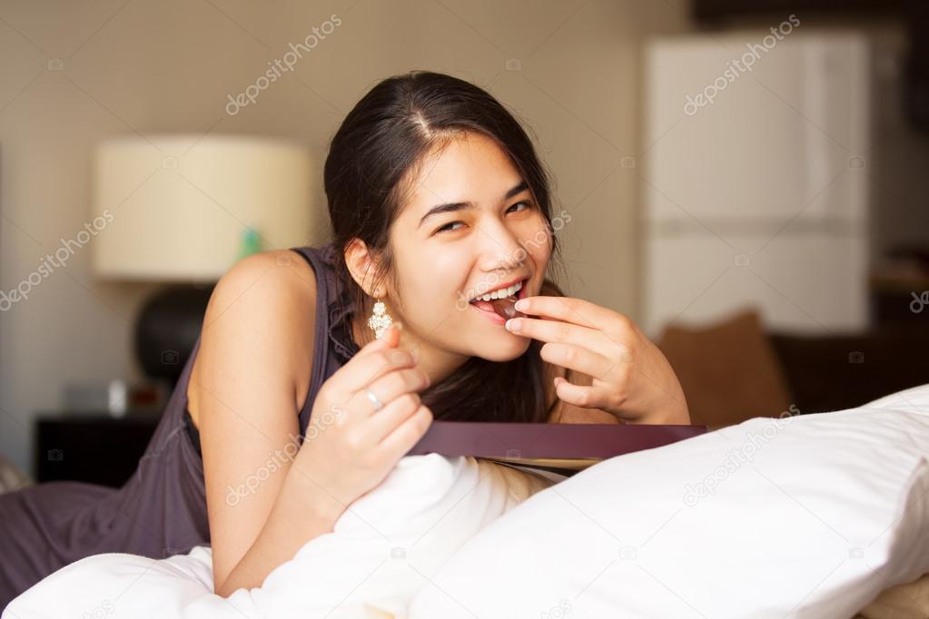 Biracial teen girl lying on bed eating chocolates, smiling