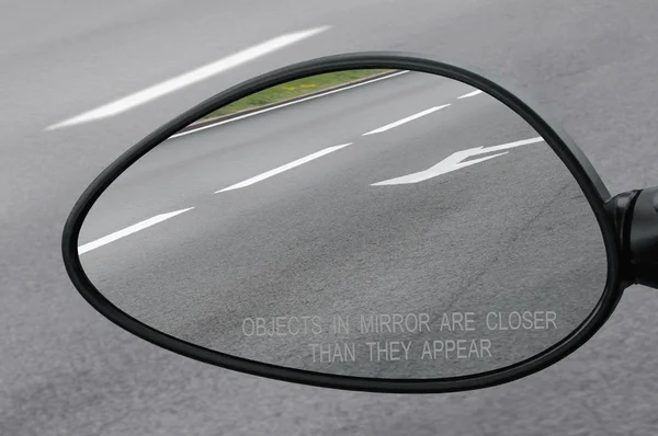 Rear view mirror with warning text objects in mirror are closer than they appear, reflecting road, left side lateral, macro closeup, tarmac asphalt background reflection, white lines, arrows marking — Stock Photo, Image