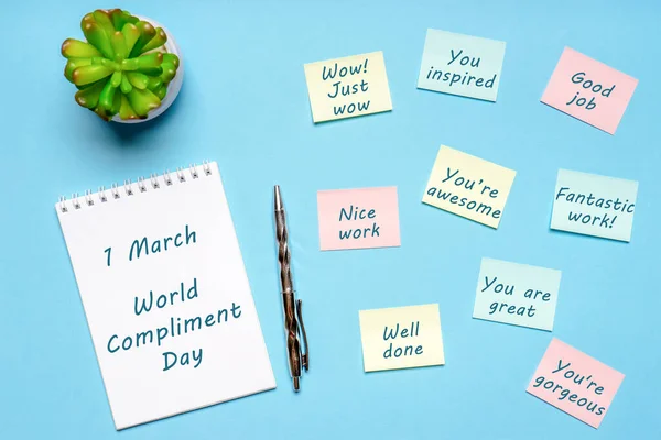 Happy World Compliment Day. Office desk with plant, notebook, pen and paper slips with compliments text for office worker such as GOOD JOB. Greeting card for world compliment day. Flat lay, top view.