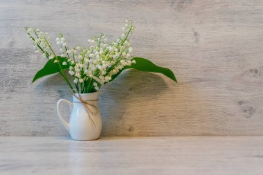 Hafif ahşap arka planda, küçük bir sürahi içinde, vadi çiçeklerinin taze zambakları. Güzel bahar natürmortu, tebrik kartı için hazırlan, boşluğu kopyala.
