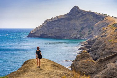 Dağlık yolda duran, deniz ve dağların güzel manzarasına hayran genç bir kadının dikiz manzarası. Genç turist kız yaz günü patikada yürüyor. Seyahat yerleri, konaklama konsepti.