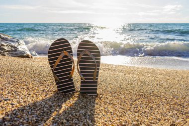 Deniz kıyısında, güneşli deniz manzarasına karşı parmak arası terlikler. Denizcilik tarzında terlikleri deniz kenarındaki küçük taşlara takla atıyor. Yaz günü deniz manzarası çok güzel. Yaz tatili, hafta sonu gezisi konsepti