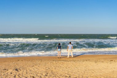 Kadın ve erkek, güneşli bir yaz gününde fırtınalı dalgalara karşı deniz kıyısındaki kumsalda dururlar. İnsanlar yaz tatilini denizde geçiriyorlar. Deniz kenarına gezi, yaz tatili konsepti..