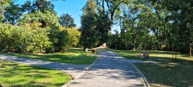 A sunny, wide-angle view captures the intersection of gravel paths and scattered park benches amidst the mature trees and lush green lawns of Rokov perivoj in Zagreb