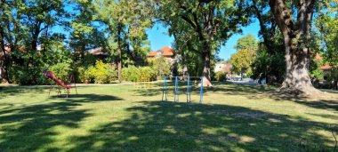 A sunny, wide-angle shot of a children's playground area with basic equipment like a slide and climbing bars, set amidst large mature trees and a grassy lawn in Rokov perivoj in Zagreb, with residential buildings visible in the background