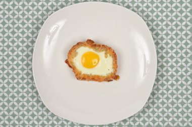 Tepeden çekilen bir çekimde, yeşil ve beyaz bir geometrik masa örtüsüne dayanan basit bir beyaz tabağa yerleştirilmiş bir tost ekmeğinin içinde pişmiş yumurta (kızarmış yumurta veya peynir kabuğuyla pişirilmiş) bir delikte tek bir yumurta yakalar.