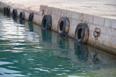 Old rubber tires hang as fenders along a stone quay above calm, turquoise harbor water in bright sunlight