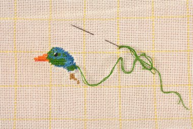 A close-up shows a partially finished needlepoint project of a small blue and green bird on canvas with two needles and green yarn
