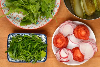 Tepeden çekimde dört kase taze sandviç ya da salata malzemesi gösteriliyor: roka, dilimlenmiş soğan, dilimlenmiş domates, turşu ve turşu, hepsi ahşap bir yüzey üzerinde.