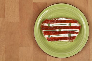 Açık yüzlü, kırmızı biberli, krem peynirli ve soğanlı bir sandviç yeşil bir tabakta servis edilir, açık ahşap arka planda geniş bir kopya alanı vardır.