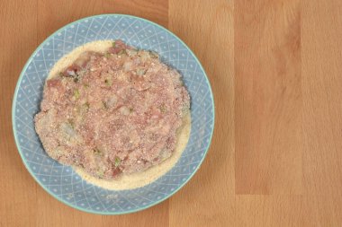 Çiğ, yassı bir et deseni mavi ve beyaz desenli bir tabakta duruyor, ekmek kırıntılarıyla kaplı, ahşap bir yüzey üzerinde dinleniyor.