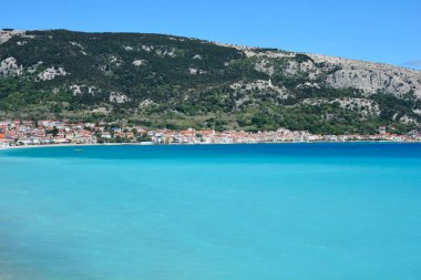 Krk Adası 'ndaki Baska şehri, berrak mavi gökyüzünün altında yeşil ve kayalık tepelerle desteklenen sakin bir turkuaz körfez boyunca kurulmuştur.
