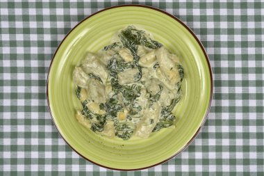 Yeşil ve beyaz kareli masa örtüsü üzerinde duran yeşil bir tabakta servis edilen kremalı ıspanak soslu gnocchi içerir.