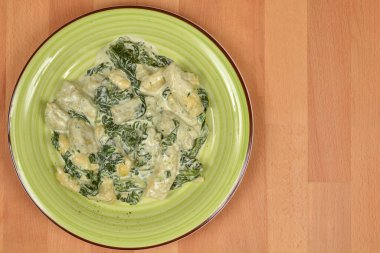 Tepeden çekimde, yeşil tabakta sunulan kremalı ıspanak soslu gnocchi görülüyor. Hafif ahşap arka planda geniş bir boşluk var.