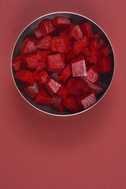 Tepedeki bir stüdyoda, koyu kırmızı arka planda kırmızı pancar kökü küpleriyle dolu bir metal kase görülüyor.