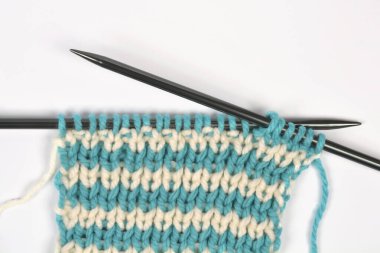 Yakın plan, beyaz arka plan üzerinde çizgili bir desen oluşturan koyu renkli iğnelerin üzerindeki mercan ve krem iplikleriyle örgü örme sürecini gösteriyor.