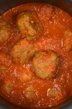 Yukarıdan aşağı, yakınlaştırılmış bir görüntü, büyük, pişmiş köftelerden oluşan bir kümeyi gösteriyor. Kalın, zengin, kırmızı bir domates sosuna batırılmış metal bir tencerenin içinde.