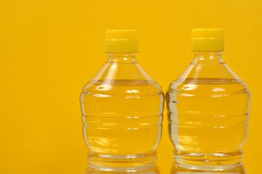 Açık sarı sıvıyla dolu iki temiz plastik şişe, muhtemelen ayçiçeği yağı, parlak sarı bir zemin üzerinde duruyor.