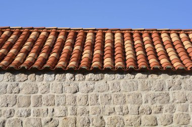 Açık mavi gökyüzünün altında, desenli, açık renkli, kaba kesim taş bloklu bir duvarın üzerinde geleneksel fayanslı bir terakotta çatısının yatay görüntüsü.