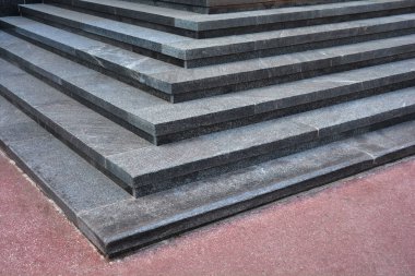 Koyu gri, pürüzlü granit basamaklardan yapılmış modern bir dış merdiven köşesinin alçak açılı yakın çekimi kırmızımsı bir zeminde duruyor.