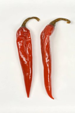İki kırmızı, parlak ve biraz buruşuk yeşil saplı kırmızı biber düz beyaz bir yüzey üzerinde dikey olarak sunulur.