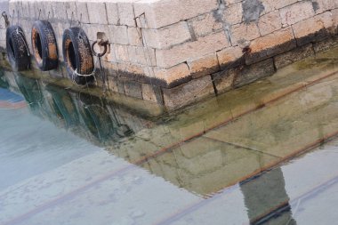 Sakin bir günde, berrak sığ deniz suyuna yansıyan siyah lastik tamponlu eski bir liman iskelesi.