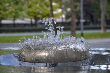 Şehir parkındaki bulanık yeşil bir arka planda modern bir metal çeşmeden fışkıran temiz suyun yakın plan görüntüsü.