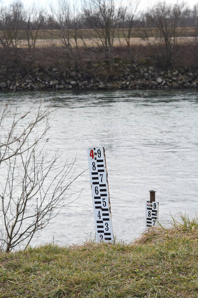 A vertical water level staff gauge stands on a grassy riverbank to measure the depth of the flowing water