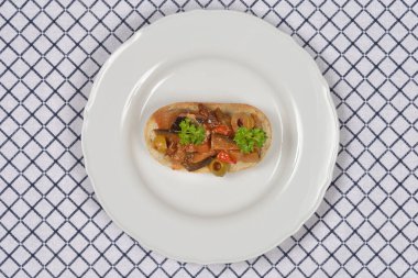 Sebzeli caponata ile kaplanmış tek bir dilim bruschetta mavi kareli masa örtüsünün üzerinde beyaz bir tabakta servis edilir.