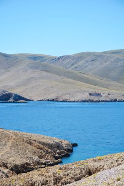 Prvic adasındaki deniz feneri Strazica Krk adasından bakıldığında açık bir gökyüzünün altında engebeli bir deniz kıyısında taş bir bina görülüyor.