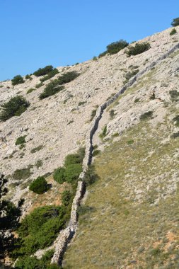 Krk adasının güney kesimindeki kayalık ve çimenli yamaçlara uzanan uzun ve geleneksel bir taş duvar berrak bir gökyüzünün altında uzanır.