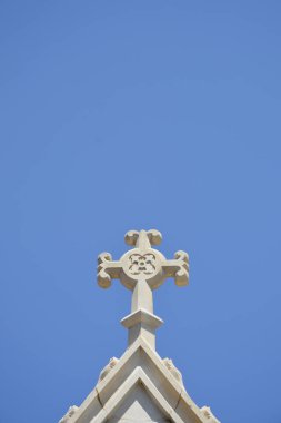 Dekoratif bir taş haç, küçük bir şapelin çatısının tepesinde berrak ve parlak mavi bir gökyüzü arka planına karşı duruyor.
